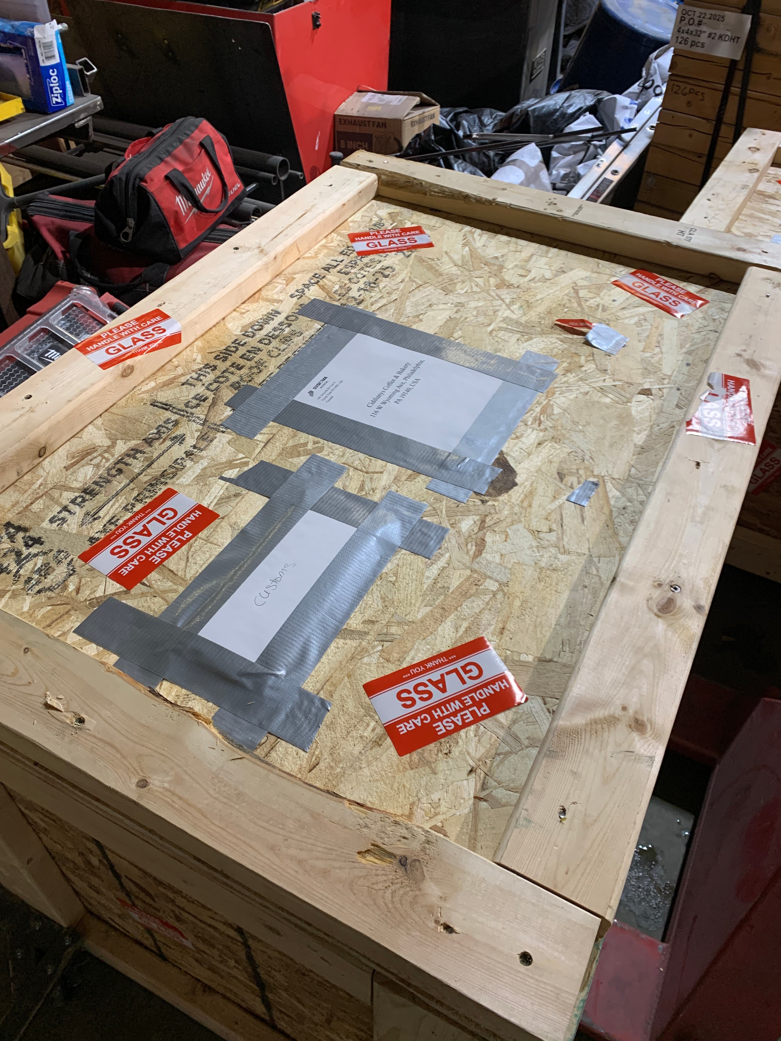 A closed crate lid marked with a taped shipping address label, customs label and red "Handle With Care — Glass" stickers.